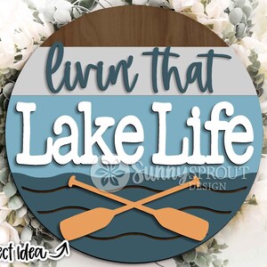Op de afbeelding: Een rond houten bord met een blauw, grijs en bruin kleurenschema. Het bord zegt "Livin' That Lake Life" met twee gekruiste roeispanen voor een golvende blauwe lijn.