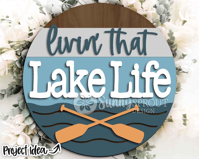Op de afbeelding: Een rond houten bord met een blauw, grijs en bruin kleurenschema. Het bord zegt "Livin' That Lake Life" met twee gekruiste roeispanen voor een golvende blauwe lijn.