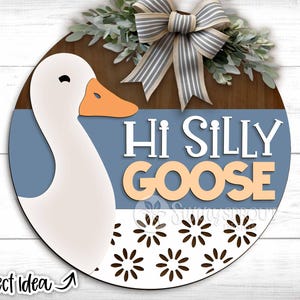 Op de afbeelding: Een rond houten bord met een witte gans en de tekst "Hi Silly Goose" in een blauw-wit bloemenpatroon.