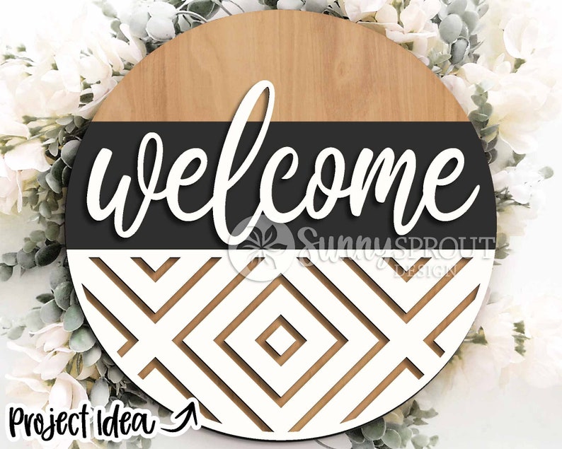 May include: A round wooden sign with a black and white geometric pattern. The top half of the sign is wood with a black band and the word "welcome" in white cursive lettering. The bottom half of the sign is a white geometric pattern with black lines.