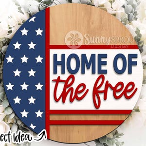May include: A round wooden sign with a red, white, and blue American flag design. The sign says "Home of the Free" in blue and red lettering.
