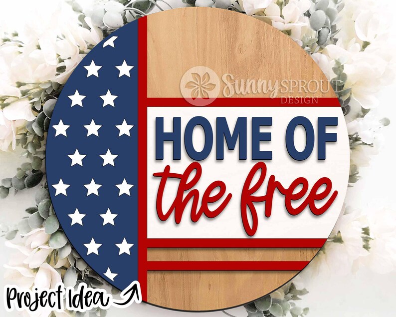 May include: A round wooden sign with a red, white, and blue American flag design. The sign says "Home of the Free" in blue and red lettering.