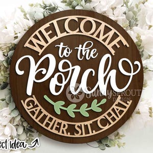 May include: A round wooden sign with the words "WELCOME to the PORCH" in white and light brown lettering. The sign also includes the words "GATHER. SIT. CHAT." and a green leaf design. The sign is surrounded by white flowers.