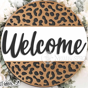 Op de afbeelding: Een rond houten bord met een luipaardprint. De bovenste helft van het bord is wit met het woord "Welkom" in zwarte cursieve letters.
