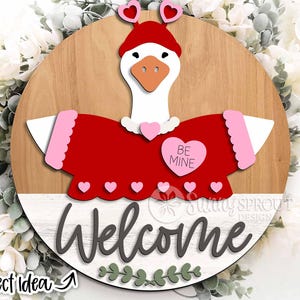 Könnte beinhalten: Ein rundes Türschild aus Holz mit einer weißen Gans, die eine rote Mütze mit herzförmigen Antennen und einen roten Pullover mit rosa Saum trägt. Ein rosa Herz auf dem Pullover trägt die Aufschrift BE MINE. Welcome steht in schwarzer Schrift.