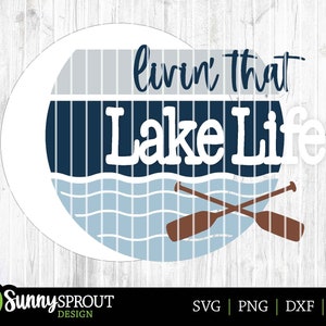 Op de afbeelding: Een witte cirkel met een blauw en grijs gestreept ontwerp en de tekst "livin' that Lake Life" in wit. Twee bruine peddels zijn gekruist over een blauw golfontwerp.
