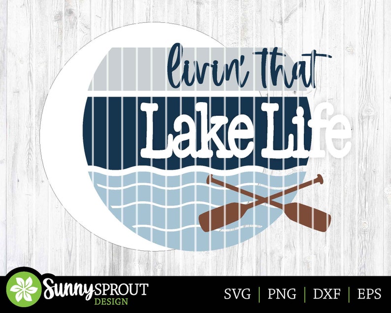 Op de afbeelding: Een witte cirkel met een blauw en grijs gestreept ontwerp en de tekst "livin' that Lake Life" in wit. Twee bruine peddels zijn gekruist over een blauw golfontwerp.