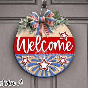 May include: A round wooden door hanger with a red, white, and blue patriotic theme. It features a bow, stars, and fireworks with the word "Welcome" in white lettering.