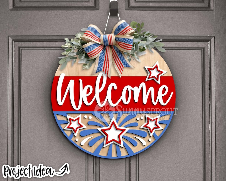 May include: A round wooden door hanger with a red, white, and blue patriotic theme. It features a bow, stars, and fireworks with the word "Welcome" in white lettering.