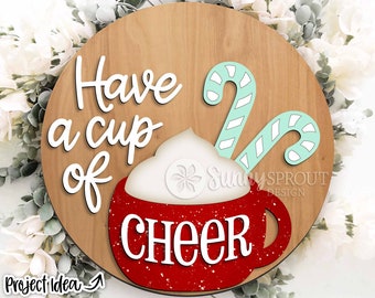 Pode incluir: Um sinal redondo de madeira com o texto "Have a cup of cheer" em letras brancas. Um copo vermelho com chantilly branco e duas bengalas de doces é retratado no sinal.