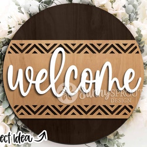 May include: A round wooden sign with a brown and light brown wood grain pattern. The sign has a black and white chevron pattern around the edge and the word "welcome" in white cursive lettering in the center.