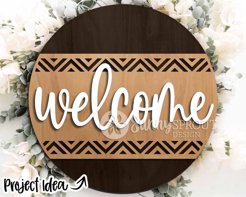 May include: A round wooden sign with a brown and light brown wood grain pattern. The sign has a black and white chevron pattern around the edge and the word "welcome" in white cursive lettering in the center.