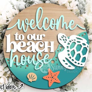 Op de afbeelding: Ronde houten bord met de tekst "welcome to our beach house" in witte en turquoise letters. Voorzien van een witte zeeschildpad, een zeeschelp en een oranje zeester. Het bord heeft een turquoise en houtnerf achtergrond.