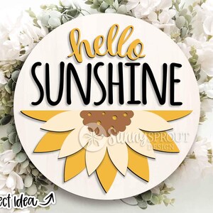 May include: A circular wooden sign featuring the words "hello SUNSHINE" in black and yellow. A sunflower design is at the bottom. The sign has a light wood grain background and is surrounded by white flowers and greenery.