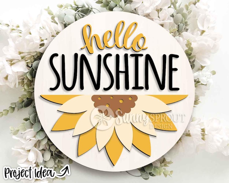 May include: A circular wooden sign featuring the words "hello SUNSHINE" in black and yellow. A sunflower design is at the bottom. The sign has a light wood grain background and is surrounded by white flowers and greenery.