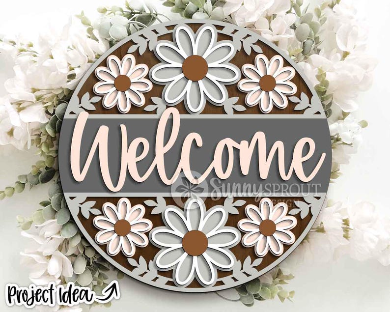 May include: A round wooden welcome sign with a floral design. The sign features the word "Welcome" in a cursive font, surrounded by daisies and decorative cutouts. The color scheme includes brown, gray, white, and light pink.