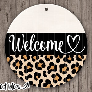 May include: Round wooden sign with a white background, a black stripe with the word "Welcome" and a heart, and a leopard print pattern.