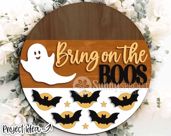 Peut inclure: Une pancarte ronde en bois avec un fond brun et orange. La pancarte dit "Bring on the Boos" en lettres jaunes et noires. Un fantôme blanc avec un visage joyeux est sur le côté gauche de la pancarte. Le bas de la pancarte a un fond blanc avec cinq chauves-souris noires sur des lunes orange.