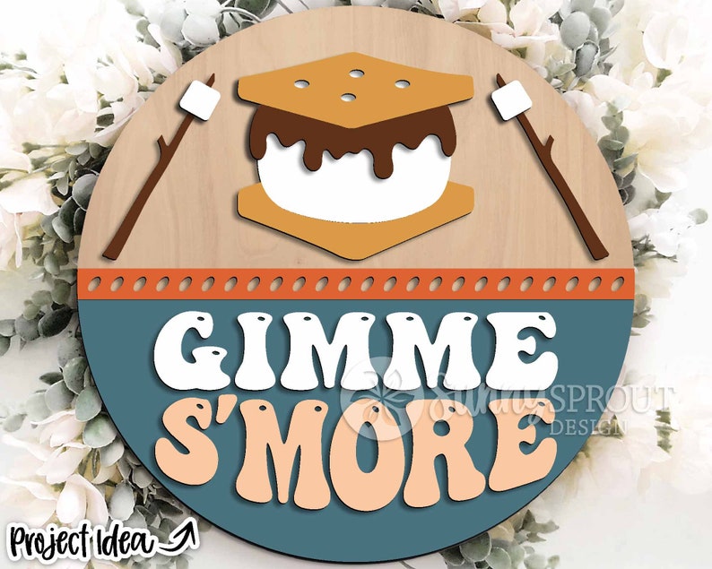 K&ouml;nnte beinhalten: Ein rundes Holzschild mit blauem Hintergrund und dem Text "Gimme S'more" in wei&szlig;en und orangen Buchstaben. Das Schild zeigt eine Grafik eines S'mores mit einem Graham-Cracker, Marshmallow und Schokolade.