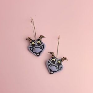 May include: A pair of dangle earrings featuring a purple cartoon dragon head with black eyes and a yellow and orange striped horn. The earrings are made of polymer clay and have gold hoops.