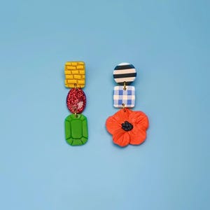 May include: A pair of colorful, whimsical earrings. One earring features a yellow brick, a red glitter oval, and a green emerald shape. The other has a striped circle, a blue and white checkered square, and an orange flower.