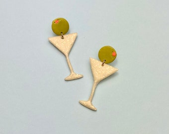 Puede incluir: Un par de pendientes de vidrio de martini de brillo blanco con dijes de aceitunas verdes. Las aceitunas tienen un pequeño detalle rosa.