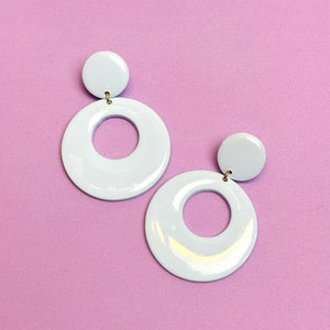 May include: A pair of white circle earrings with a smaller circle cut out in the center. The earrings are on a light pink background.