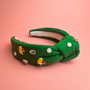 以下が含まれることがあります： 白と黄色の装飾が施された緑色の結び目のヘッドバンド。ヘッドバンドはラインストーンと小さな黄色と赤のフィギュアで飾られています。