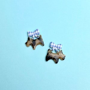 Peut inclure: Une paire de boucles d'oreilles en forme de chien brun et noir avec des nœuds en vichy bleu et blanc.