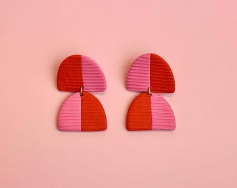 Puede incluir: Un par de pendientes geométricos de color rosa y rojo. Los pendientes están hechos de arcilla y tienen forma de semicírculo.