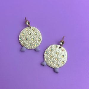 May include: A pair of white and gold earrings with a geometric pattern and iridescent accents. The earrings have a round shape and three small purple feet.