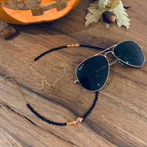May include: A black beaded glasses chain with orange and clear beads. The chain is draped over a pair of black aviator sunglasses with gold frames. The sunglasses are resting on a wooden surface. A carved pumpkin is visible in the background.