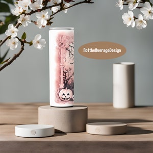 May include: A white tumbler with a pink and white illustration of a spooky Halloween scene with a jack-o-lantern, trees, and a building. The text "NottheaverageDesign" is visible in the background.