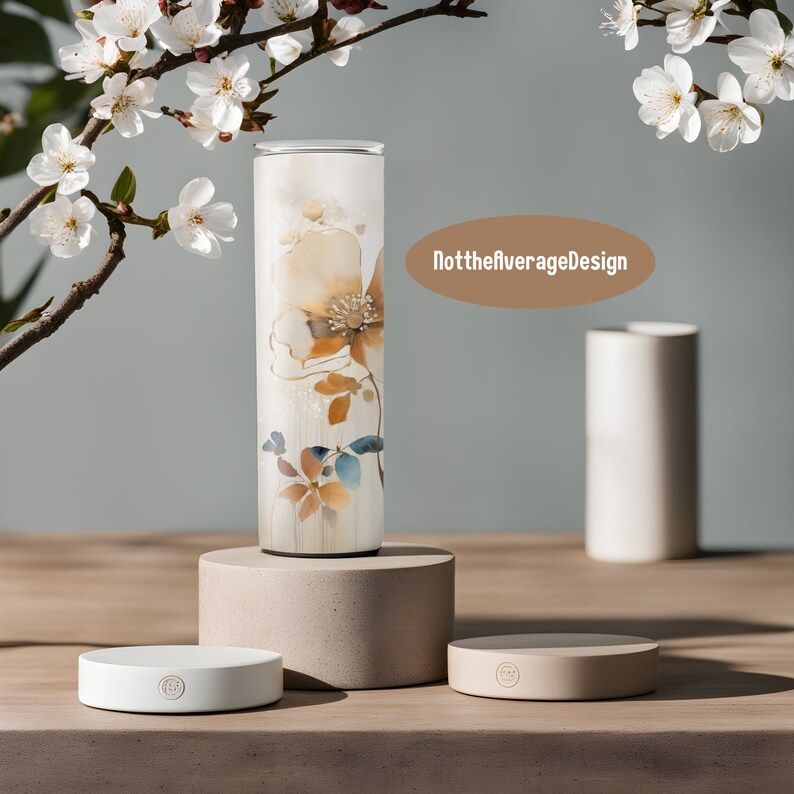 May include: A white tumbler with a floral design in shades of brown, yellow, and blue. The tumbler is sitting on a beige pedestal. The text "NottheaverageDesign" is visible in the background.