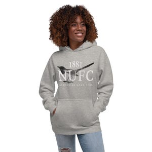 May include: A light gray hoodie with a front pocket and a drawstring hood. The hoodie features a black and white magpie graphic with the text "1881 NUFC Newcastle Upon Tyne."