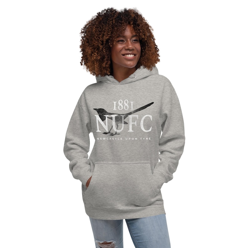 May include: A light gray hoodie with a front pocket and a drawstring hood. The hoodie features a black and white magpie graphic with the text "1881 NUFC Newcastle Upon Tyne."