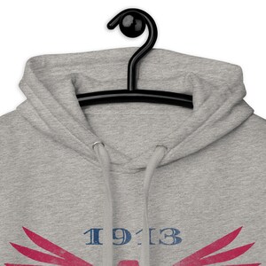 Może przedstawiać: Szara bluza z kapturem z niebieskim nadrukiem "1913" i czerwono-r&oacute;żowym wzorem. Bluza wisi na czarnym wieszaku.