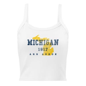 Pode incluir: Um top curto sem mangas branco com um mapa do Michigan em estilo aguarela amarelo. O texto MICHIGAN está impresso em azul escuro, seguido pelo ano 1817 e pela localização ANN ARBOR numa fonte sem serifa azul escura a condizer.