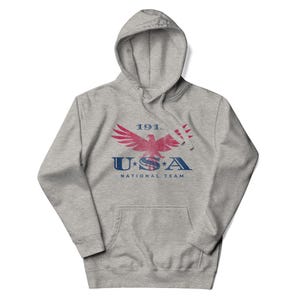 Może przedstawiać: Szara bluza z kapturem z przednią kieszenią i sznurkiem. Z przodu znajduje się czerwono-niebieska grafika orła z napisem "1911 USA NATIONAL TEAM".