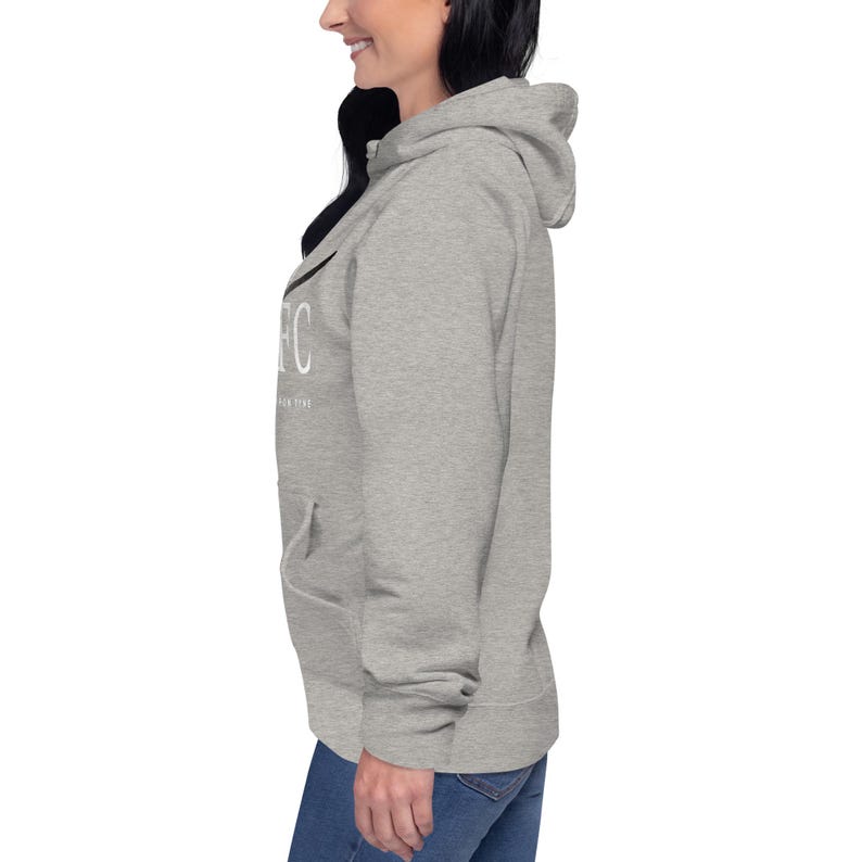 May include: A light gray hoodie with a drawstring hood and a front pocket. The hoodie features white text on the chest, including the letter "C". The model is wearing blue jeans.