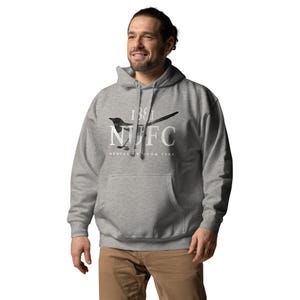 May include: A gray hoodie with a front pocket and a drawstring hood. The hoodie features a black and white graphic of a bird and the text "1881 NUFC NEWCASTLE UPON TYNE" in white. The person is wearing brown pants.
