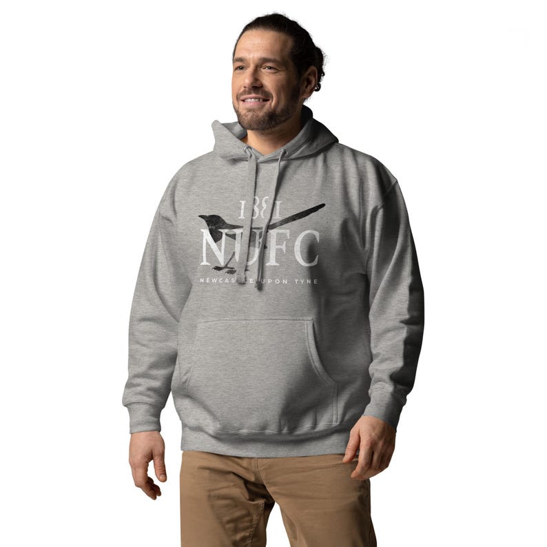 May include: A gray hoodie with a front pocket and a drawstring hood. The hoodie features a black and white graphic of a bird and the text "1881 NUFC NEWCASTLE UPON TYNE" in white. The person is wearing brown pants.