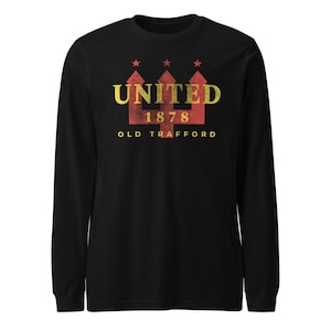 May include: A black long-sleeve shirt featuring a graphic with the word UNITED in gold, the number 1878, and the text OLD TRAFFORD. The design includes a red trident shape with three small red stars positioned above the word UNITED.