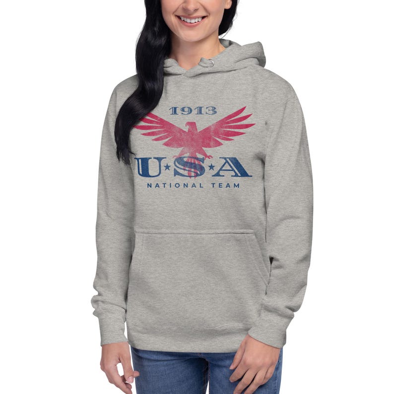 Może przedstawiać: Jasnoszara bluza z kapturem z przednią kieszenią i sznurkiem. Z przodu znajduje się czerwono-niebieska grafika z napisem "1913 USA NATIONAL TEAM" i wzorem orła.