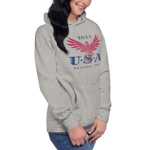 Może przedstawiać: Szara bluza z kapturem z przodu z grafiką przedstawiającą czerwonego orła, napis "1913" oraz słowa "USA National Team" w kolorze niebieskim. Bluza posiada kaptur ze sznurkiem i przednią kieszeń.