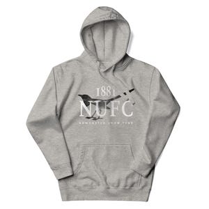 May include: A gray hooded sweatshirt with a front pocket. The sweatshirt features a black and white magpie design with the text "1881 NUFC NEWCASTLE UPON TYNE". The hoodie is made of a soft material.