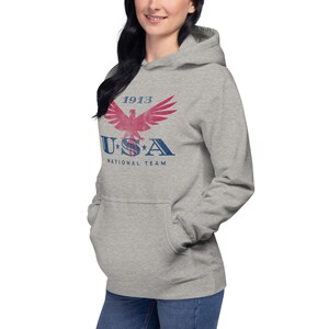 Może przedstawiać: Jasnoszara bluza z kapturem z nadrukiem z przodu przedstawiającym czerwono-niebieski wz&oacute;r orła z napisem "1913 USA National Team". Bluza z kapturem ma kaptur ze sznurkiem i przednią kieszeń.