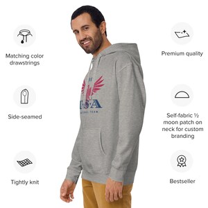 Może przedstawiać: Jasnoszara bluza z kapturem z przednią kieszenią i kapturem. Bluza z kapturem ma napis "USA National Team" z grafiką orła. Ma sznurki w dopasowanym kolorze i jest ciasno dziana. Bluza jest szyta z boku.
