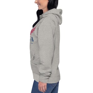 Może przedstawiać: Jasnoszara bluza z kapturem z przednią kieszenią i kapturem. Bluza z kapturem ma czerwono-niebieską grafikę na lewej piersi z napisem "USA Team". Modelka ma na sobie niebieskie dżinsy. Bluza z kapturem jest wykonana z miękkiego, wygodnego materiału.