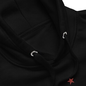 May include: Close-up of a black hooded sweatshirt. The hood features a drawstring with metal eyelets. A small red star is visible near the bottom. The sweatshirt is made of a soft, comfortable material.